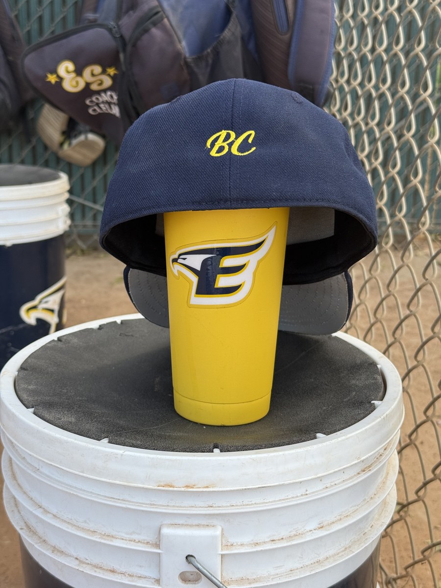 A navy El Segundo Eagles cap with Belynda's initials, BC, embroidered in yellow script on the side, resting on an Eagles cup on a ball bucket at the field