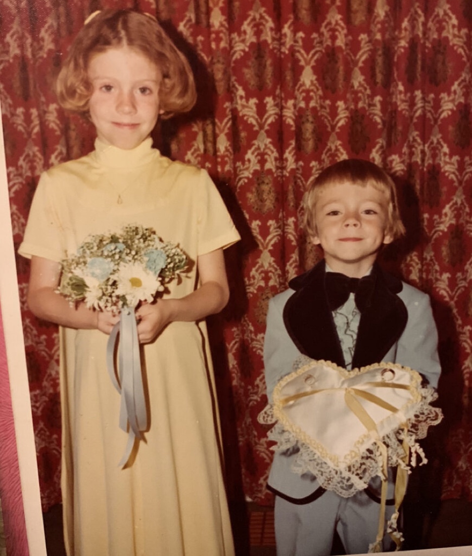 Belynda and Steve as children at a wedding, she in a long yellow dress holding a bouquet, he in a pale blue suit holding the ring pillow